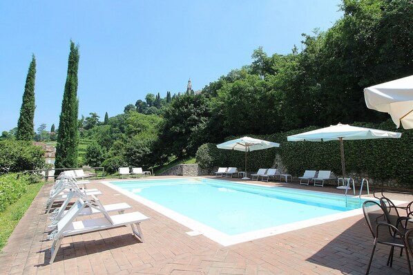 A rectangular swimming pool with a brick border, surrounded by lounge chairs and umbrellas, set against a backdrop of trees and a hill.