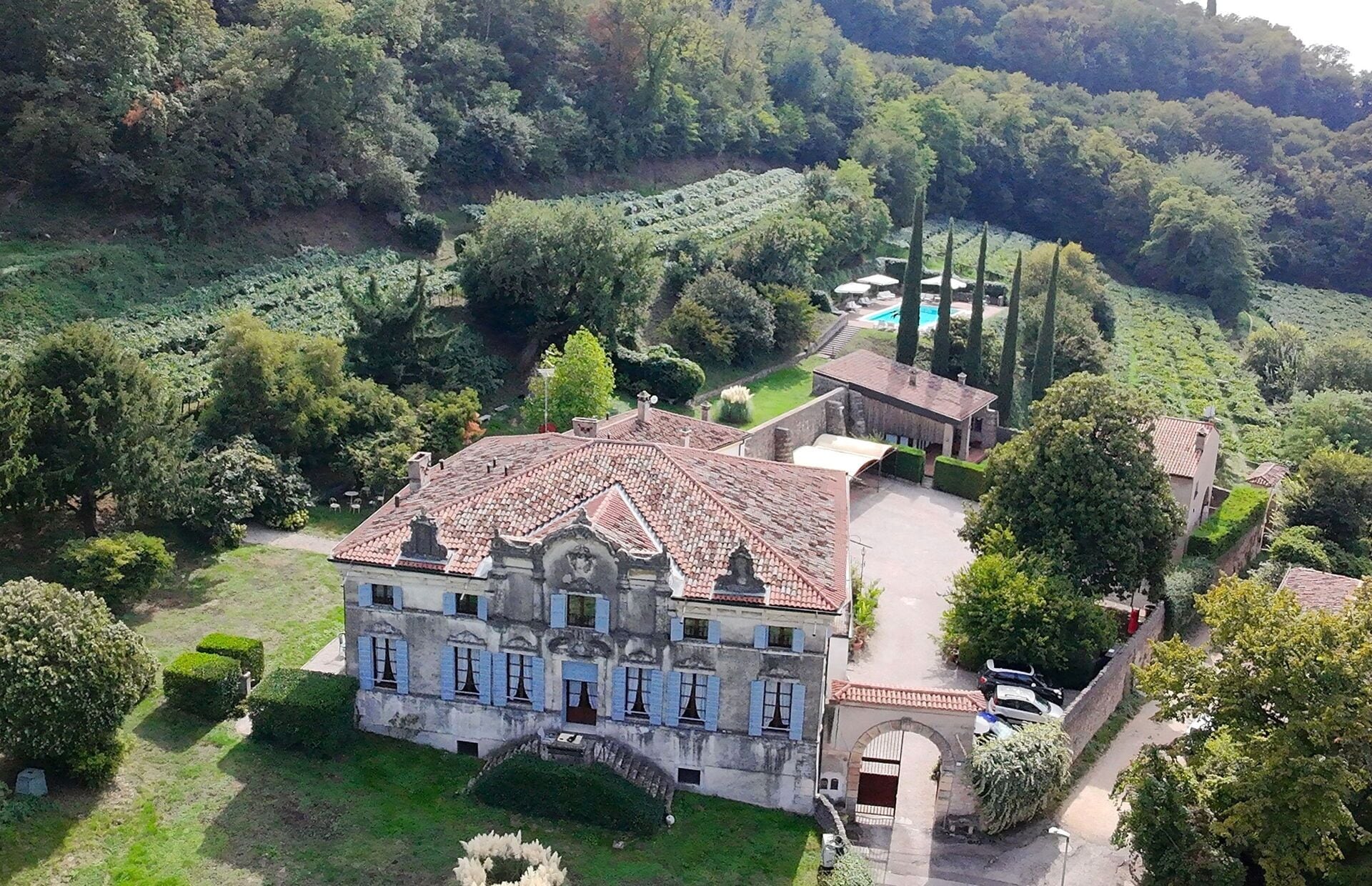 A large, historic building with a tiled roof, surrounded by lush greenery and a swimming pool.