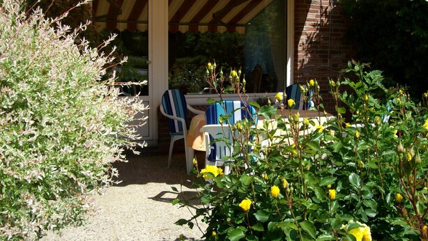 Une terrasse couverte avec une table et des chaises, un bâtiment en briques et un jardin avec des fleurs jaunes.