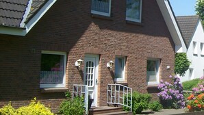 Une maison à deux étages, construite en briques, avec un toit en pente, un porche à l’avant et un petit jardin fleuri.