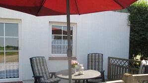 Une terrasse extérieure avec une table ronde, deux chaises, un parasol rouge et une vue sur un bâtiment blanc doté de fenêtres.