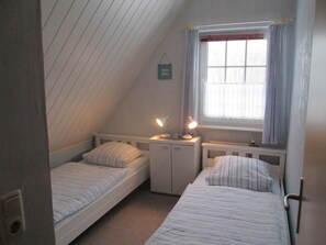 Une petite chambre à coucher composée d’une seule pièce, avec deux lits, une fenêtre avec des rideaux, un radiateur et un petit bureau avec une lampe.