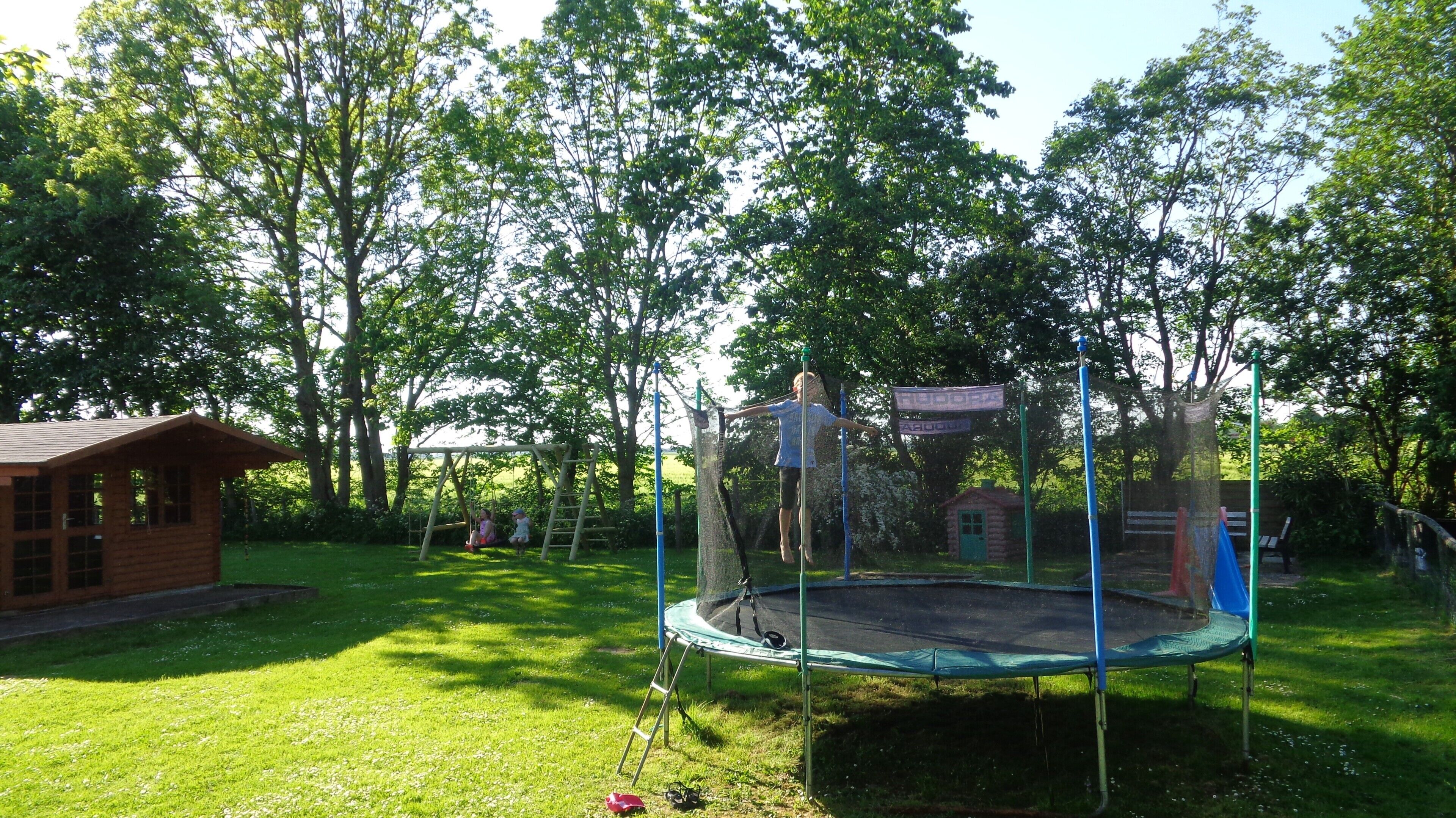 A wooden cabin, a trampoline, and a swing set in a grassy yard with trees.