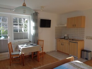 Cocina compacta con armarios de madera, una mesa de comedor con sillas, un televisor montado en la pared y una ventana con persianas.