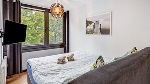 A bedroom with a bed, a gray sofa, a TV, a window with curtains, and a wall-mounted picture.