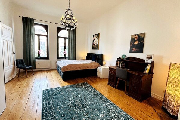 A bedroom with a bed, a desk, a chair, a chandelier, and artwork on the walls.