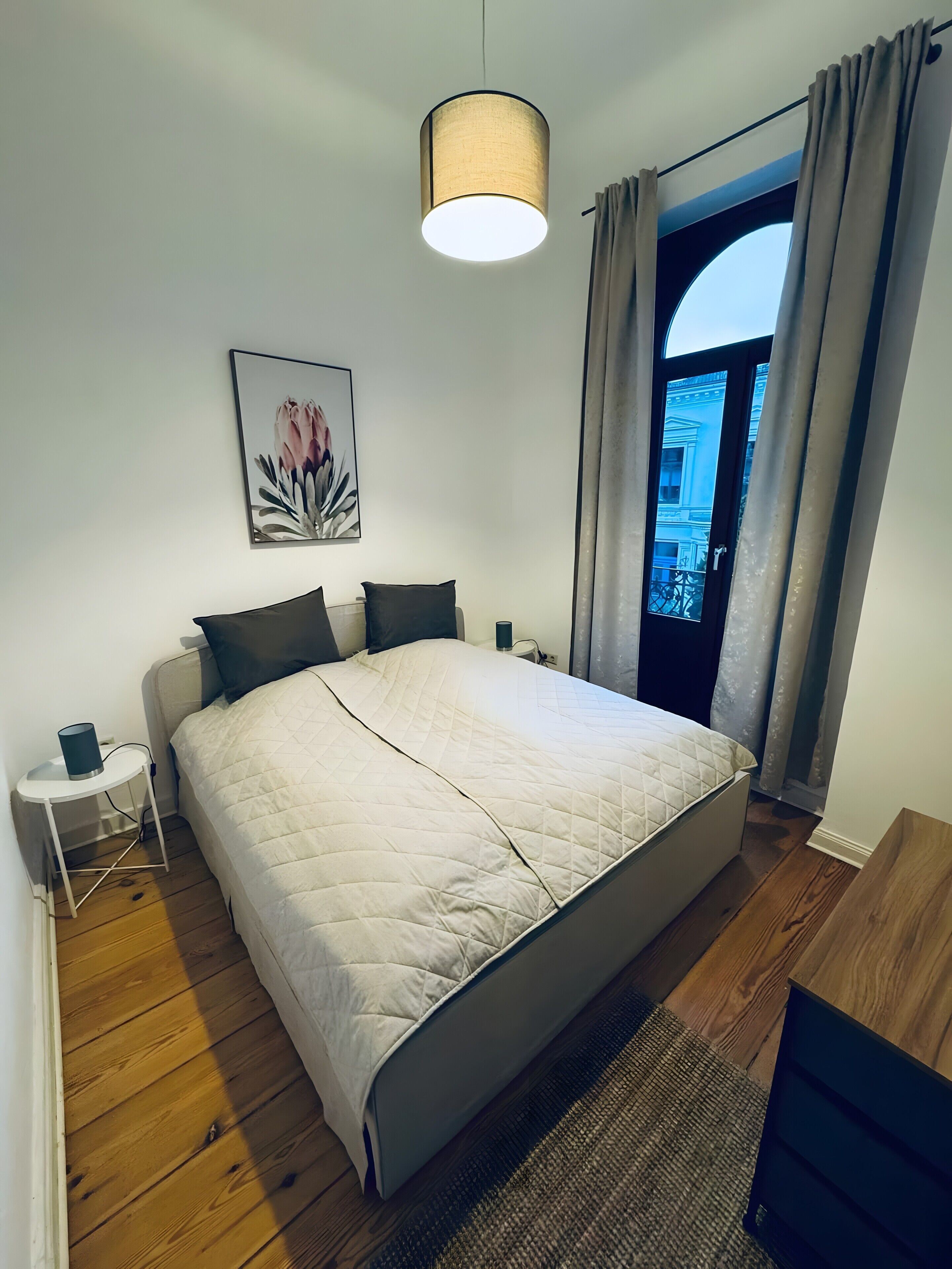 Un dormitorio con una cama, una mesita de noche, una lámpara, un cuadro y una ventana con cortinas.