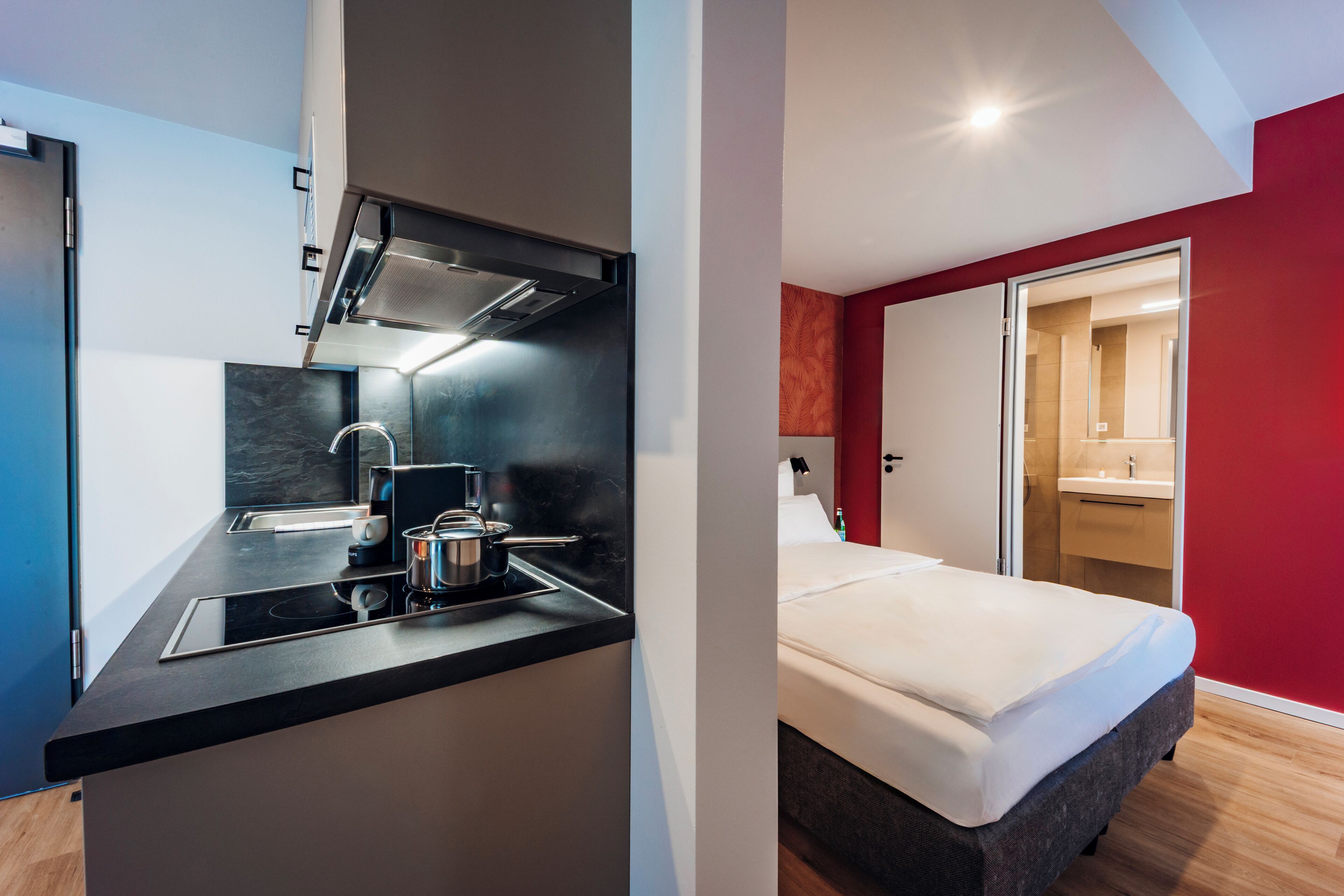 Cocina moderna con encimera negra y electrodomésticos de acero inoxidable, comedor independiente y un dormitorio con cama grande y paredes rojas.