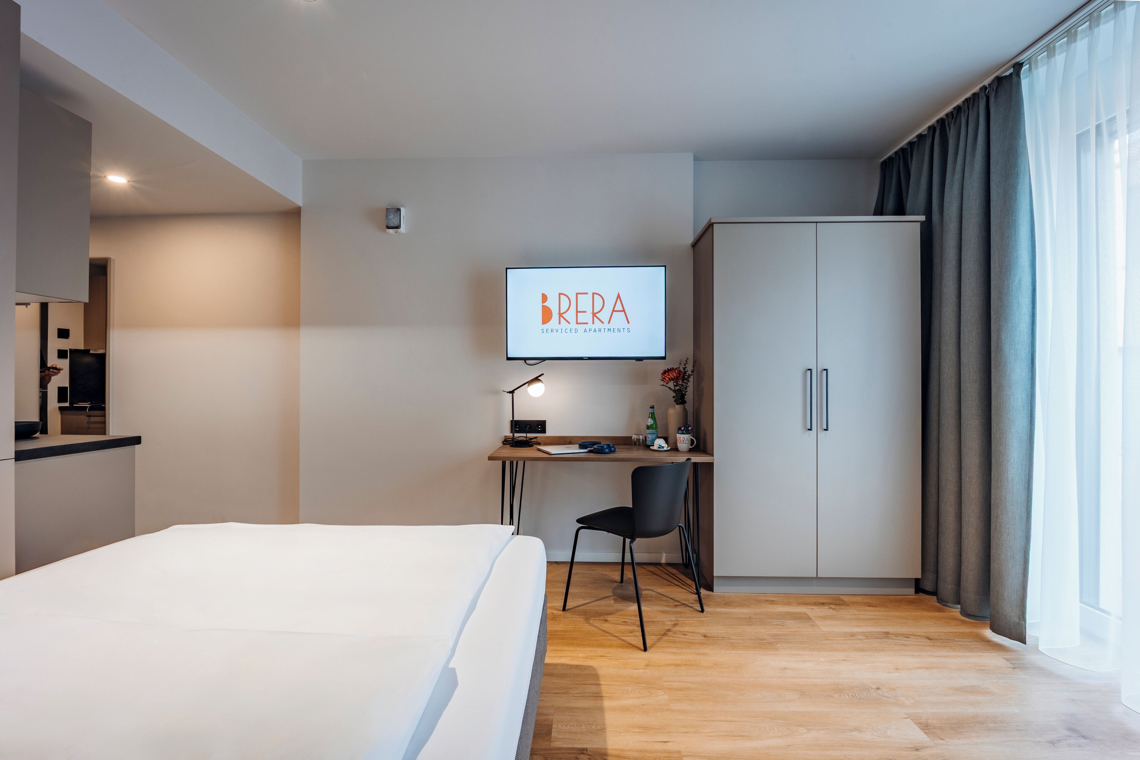 A modern hotel room with a bed, a desk with a computer and lamp, a TV displaying 'BRERA', and a wardrobe.