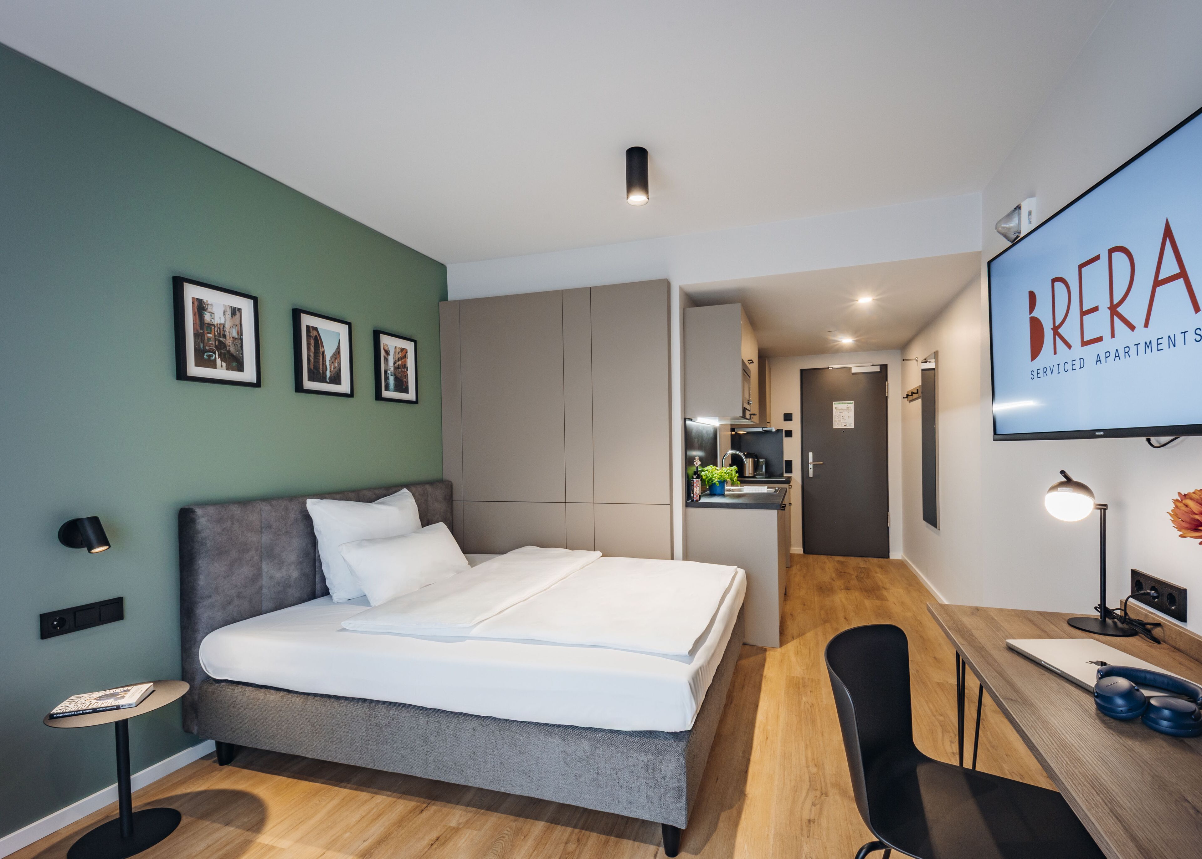 Een moderne slaapkamer met een bed, nachtkastje en een bureau met stoel. Achterin is een kitchenette te zien.