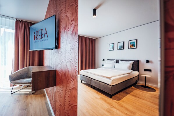 Un dormitorio moderno con cama, escritorio y televisión mostrando “BRERA SERVICED APARTMENTS”.