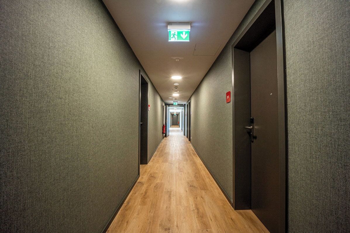 A long, narrow hallway with wooden flooring and grey walls, leading to a door at the end.