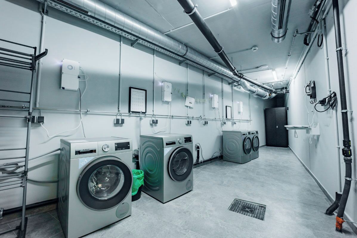 A laundry facility with multiple washing machines and dryers, ventilation ducts, and a metal rack for hanging clothes.
