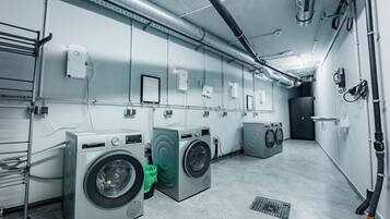A laundry facility with multiple washing machines and dryers, ventilation ducts, and a metal rack for hanging clothes.