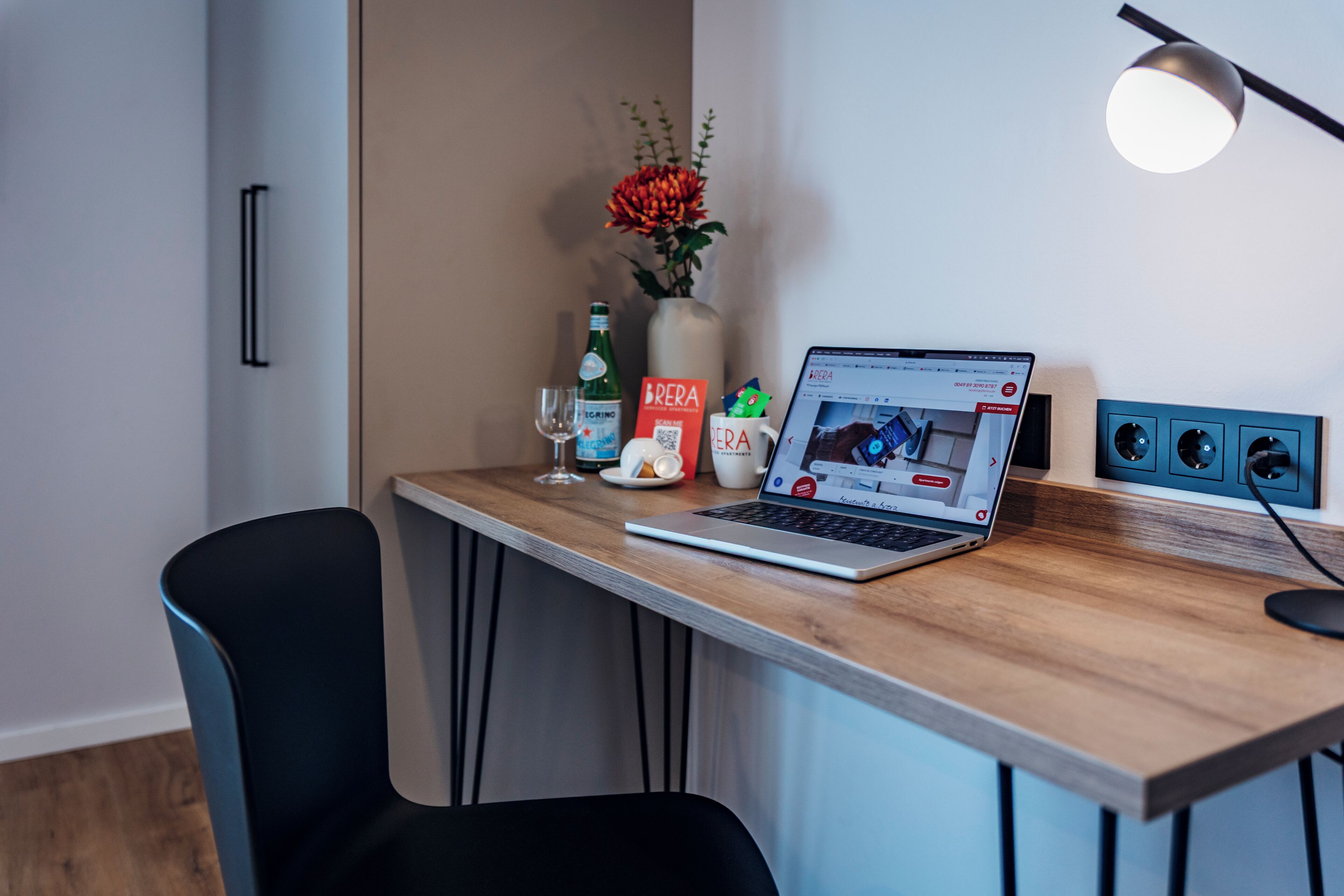 A modern workspace with a laptop, a vase with flowers, a bottle, and a mug on a wooden desk.
