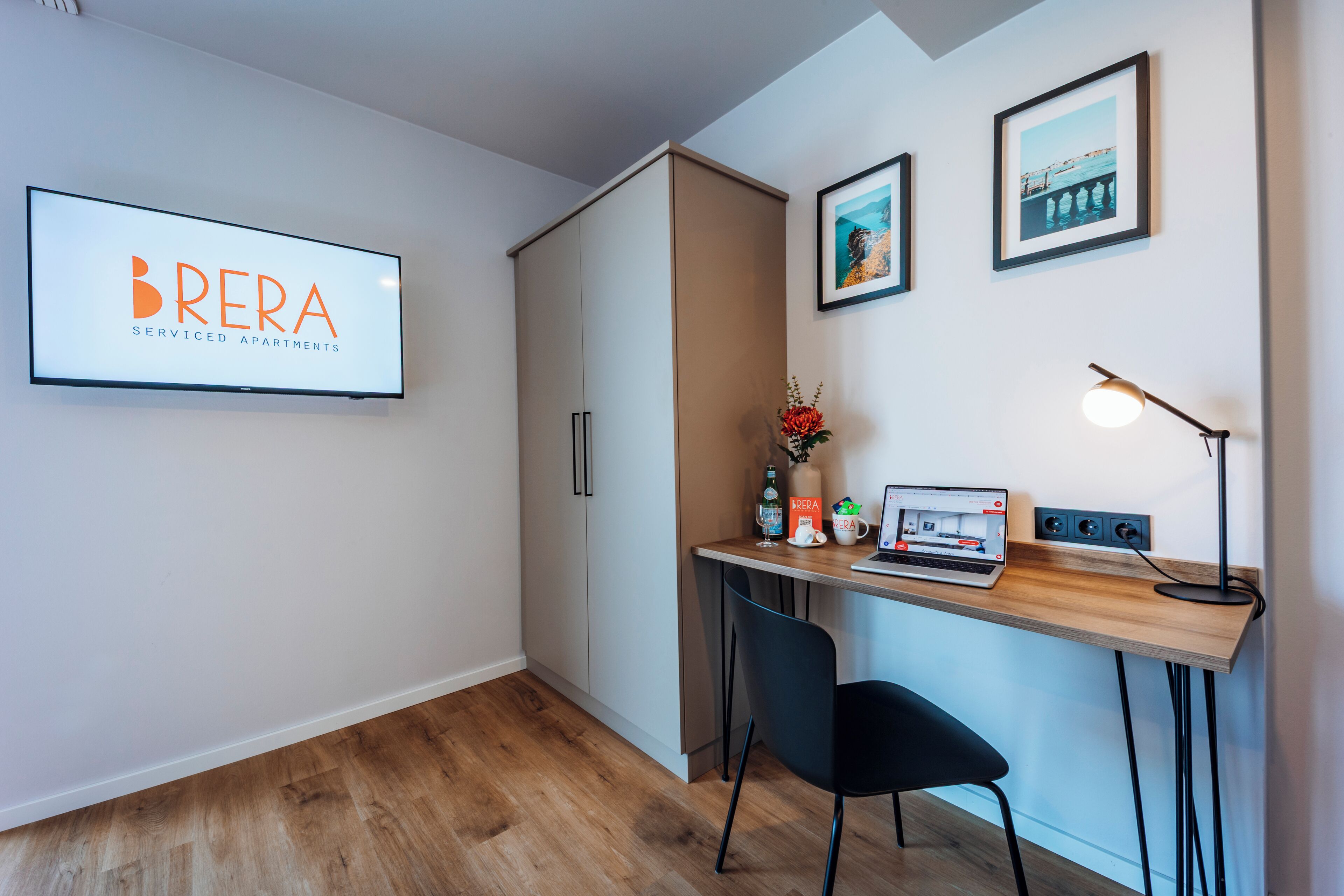 Un espace de bureau moderne comprenant un bureau, une chaise et un ordinateur. On y trouve également un tableau encadré accroché au mur, une lampe et un vase rempli de fleurs.