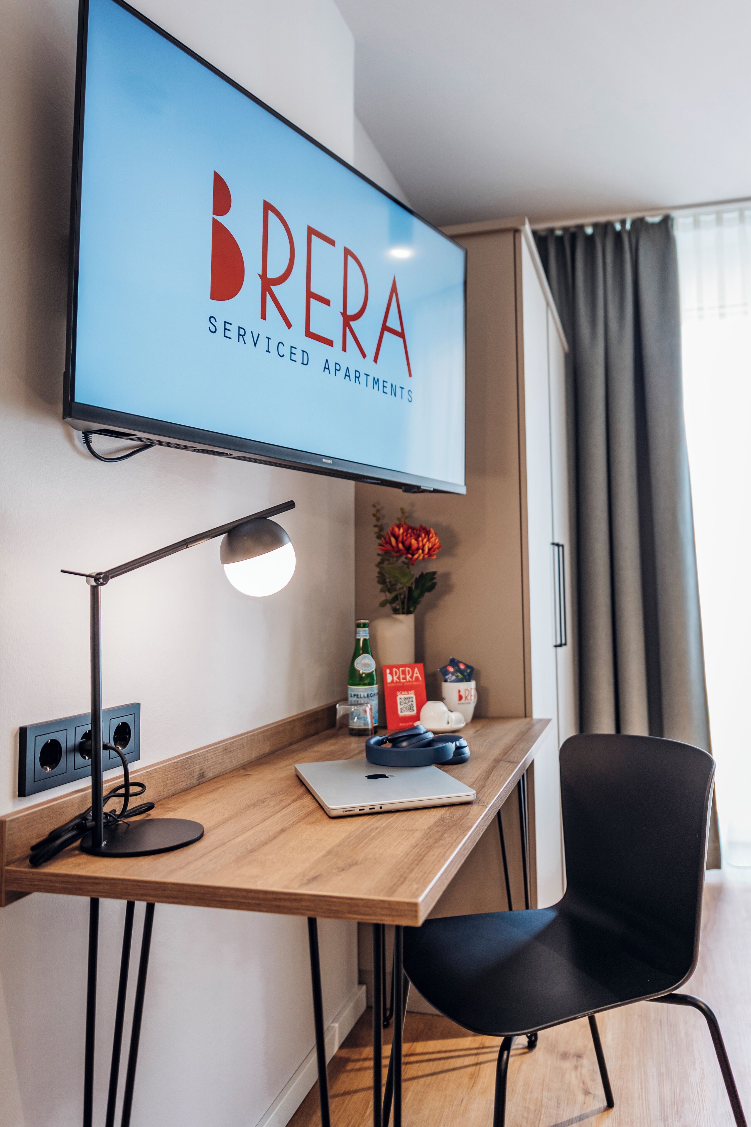 Une chambre moderne avec un bureau en bois, une chaise noire, un téléviseur fixé au mur qui affiche le logo « BRERA Serviced Apartments », une lampe, une bouteille et un vase avec des fleurs.