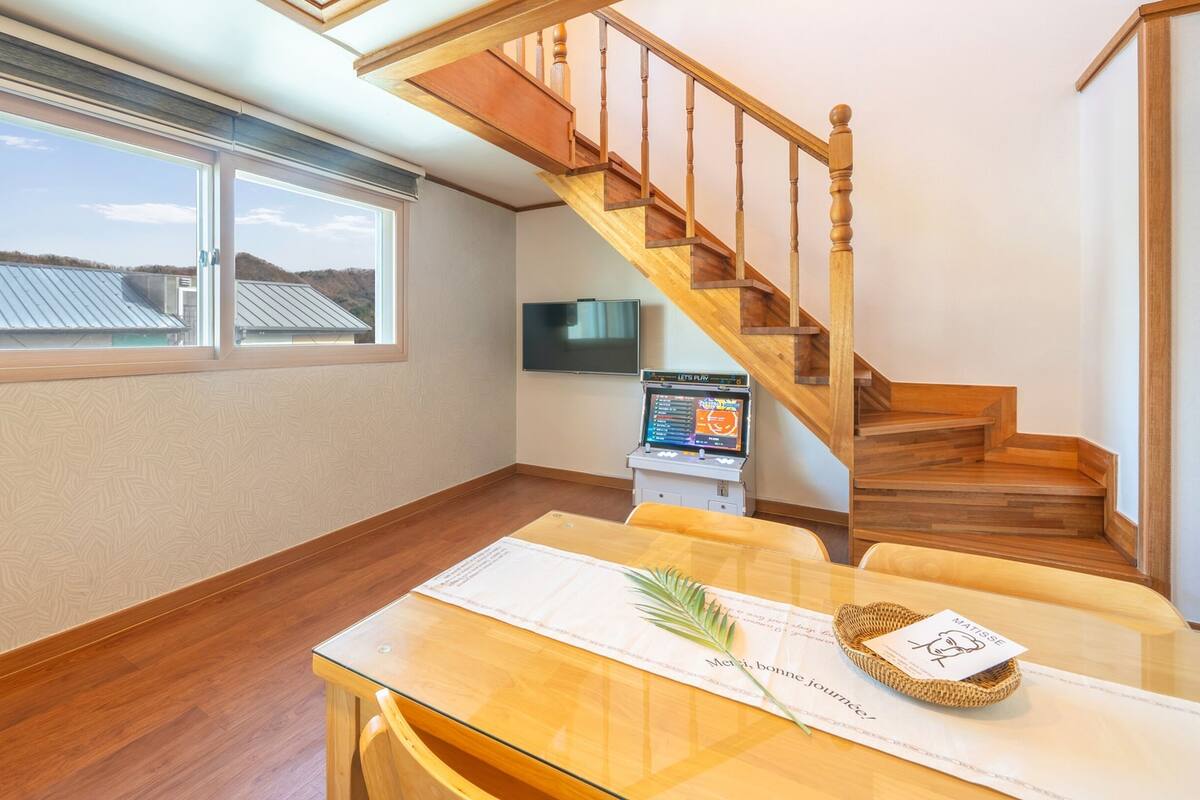 Een ruime kamer met een houten trap, een eethoek met een tafel en stoelen, een aan de muur bevestigde televisie, en grote ramen met uitzicht op het dak en het landschap.