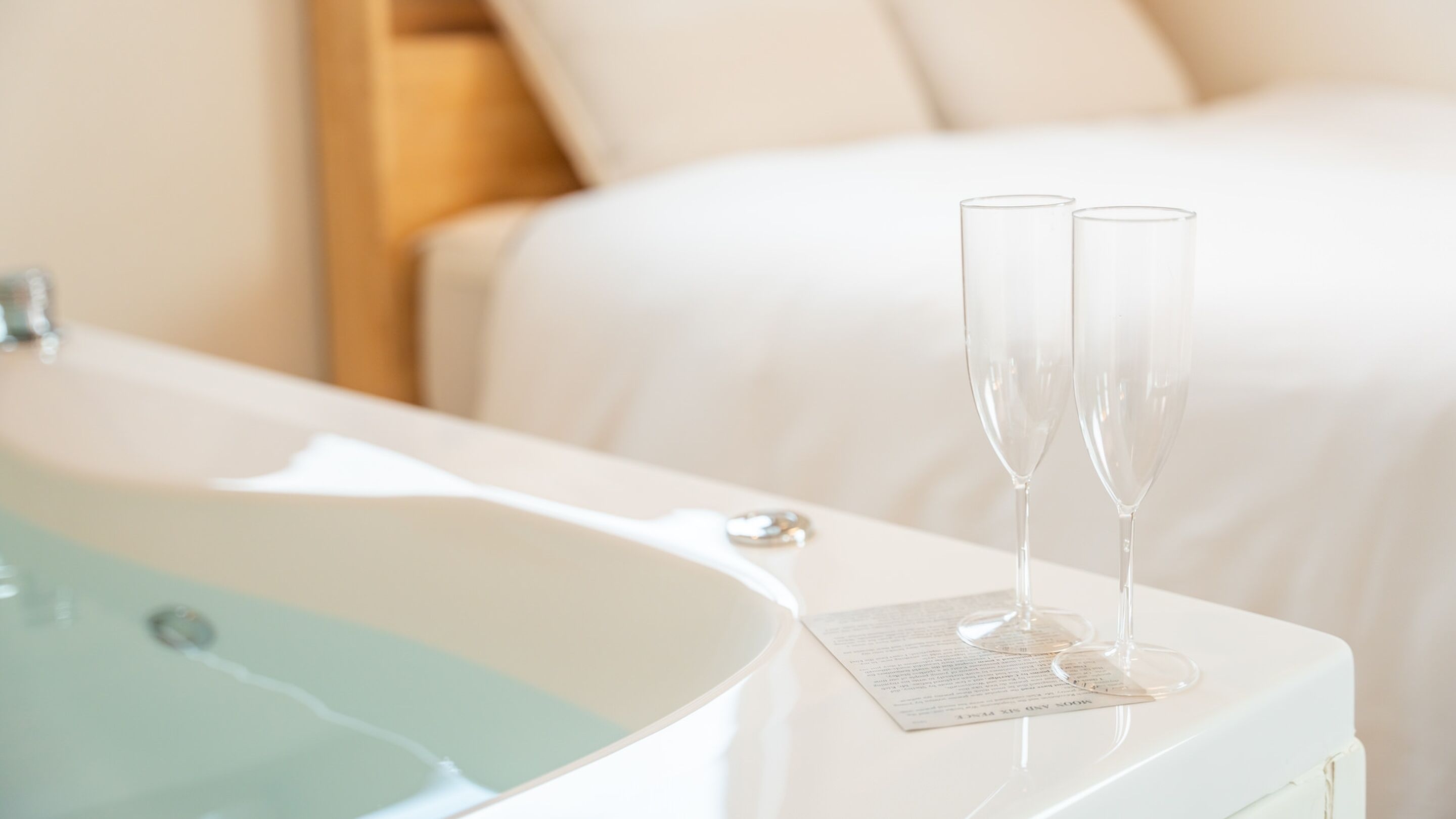 A white bathtub with two clear glasses on a white tray, a white bed, and a white wall.
