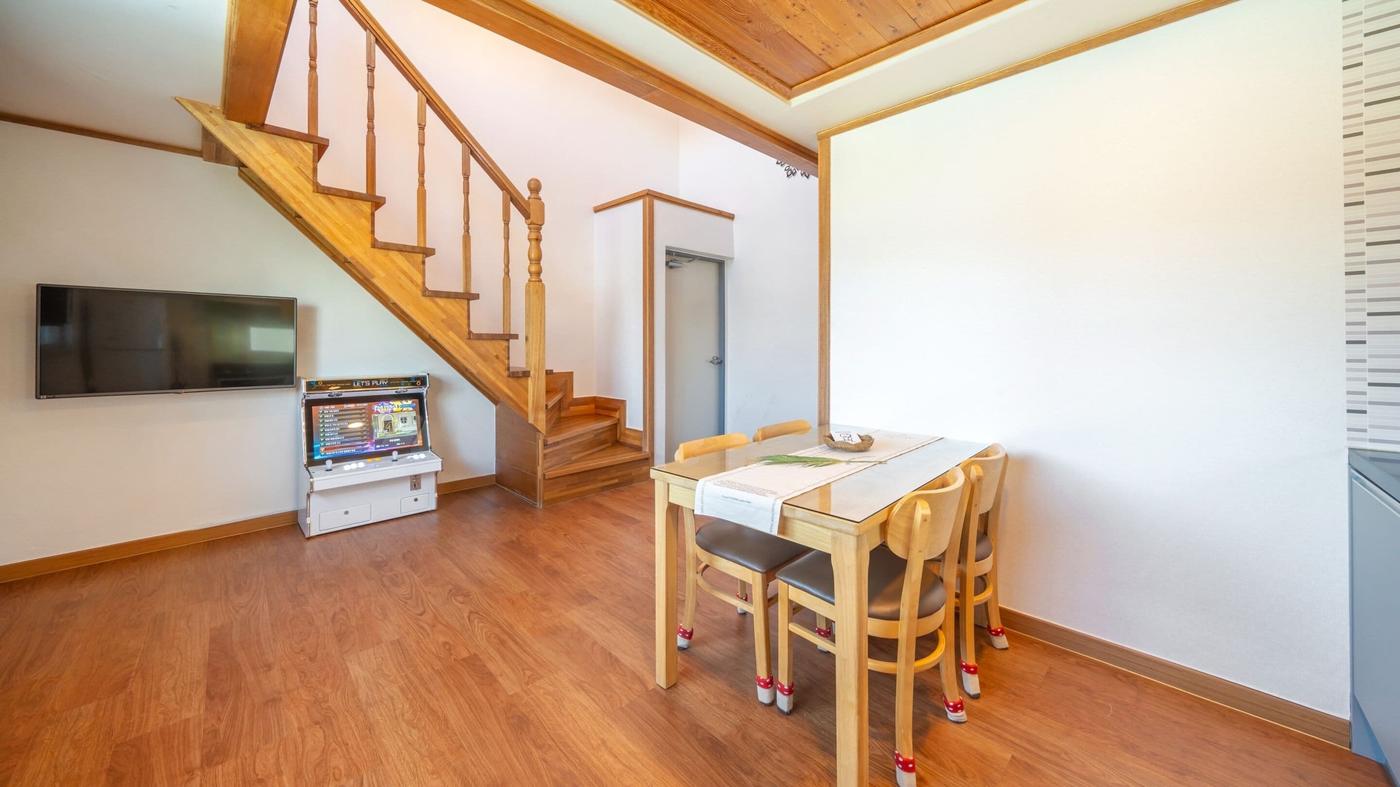 Una habitación con una escalera de madera, un televisor de pantalla plana, una máquina de karaoke y una mesa de comedor con cubierta de vidrio y sillas.