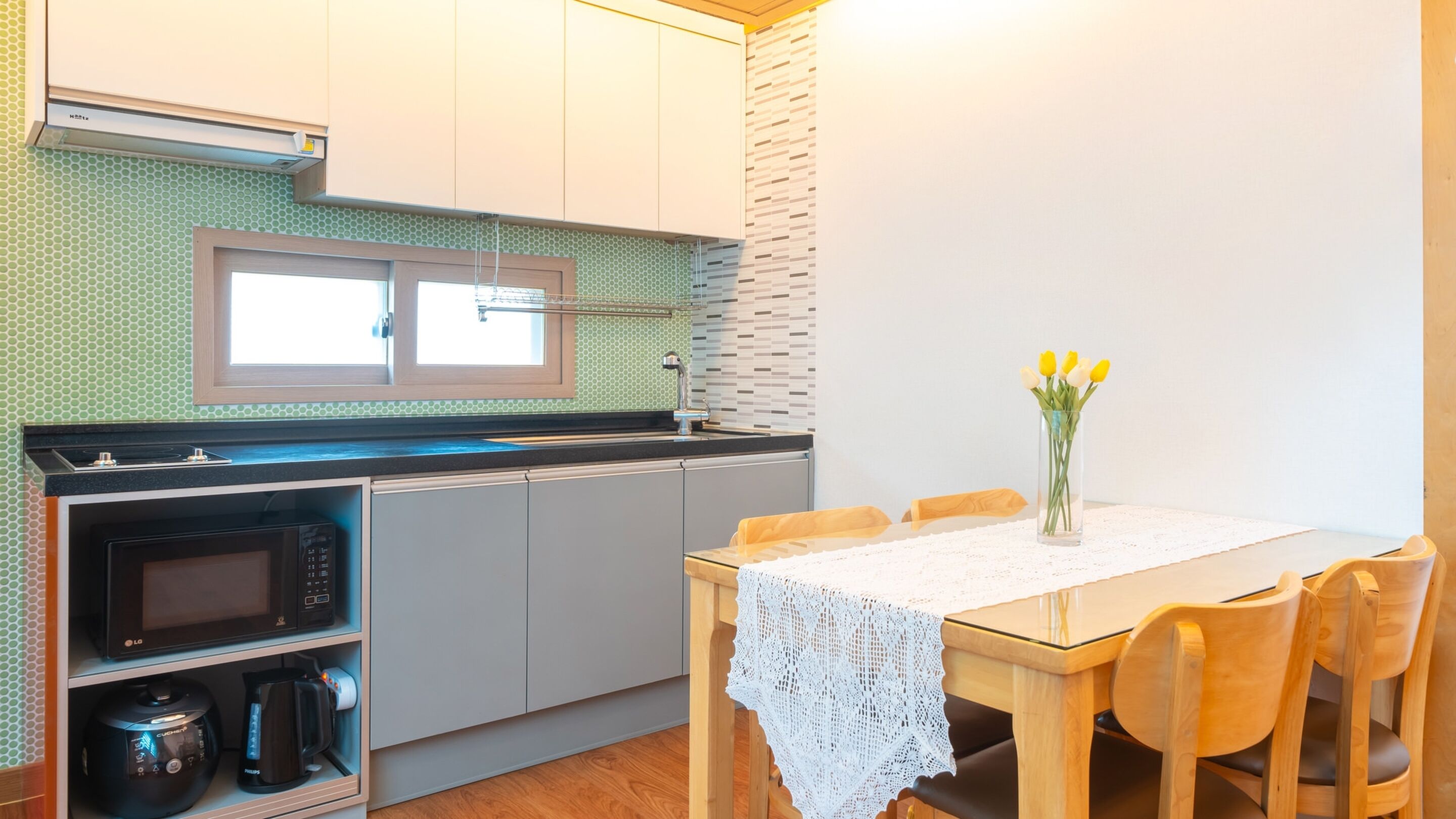Eine kompakte Küche mit Mikrowelle, Backofen und Kaffeemaschine links, einem kleinen Fenster über dem Spülbecken und einem Esstisch mit Stühlen rechts.