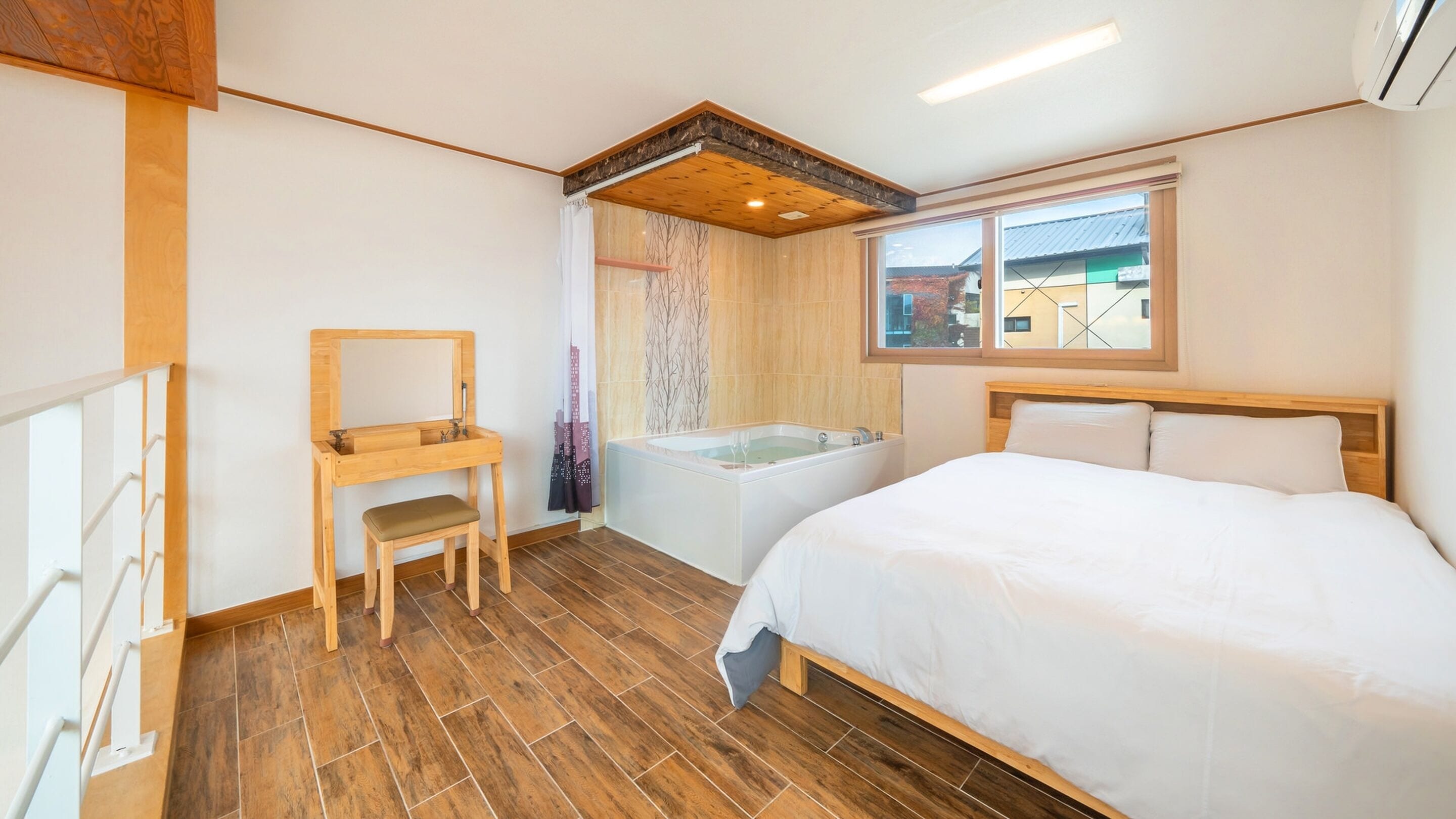 A bedroom with a wooden bed, a wooden desk with a chair, a bathtub, and a window with a view of buildings.
