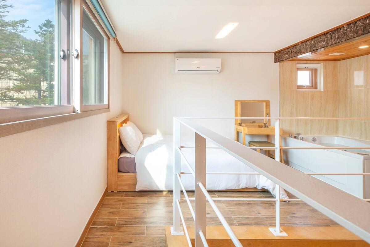 Een slaapkamer met een houten vloer, een bed met witte beddengoed, een raam met uitzicht op bomen, en een badkamer met een bad en wastafel.
