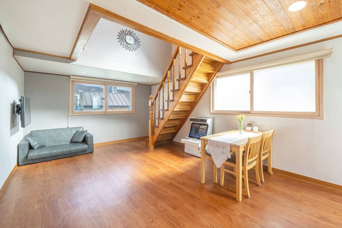 Een ruime woonruimte met een houten trap, een bank, een eettafel met stoelen en een groot raam dat natuurlijk licht binnenlaat.