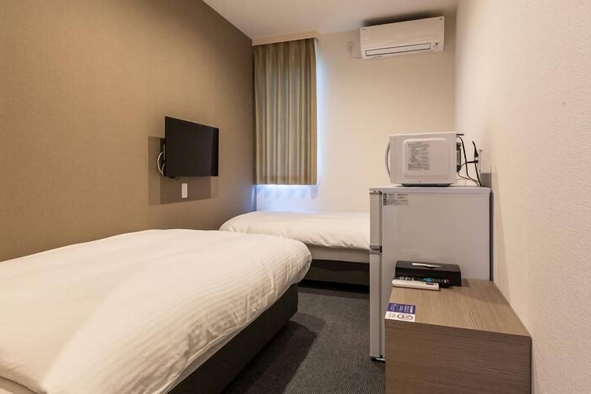 A hotel room with two single beds, a TV mounted on the wall, an air conditioning unit, and a small desk with a printer.