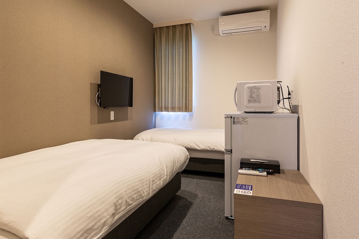 A hotel room with two beds, a television, an air conditioner, and a refrigerator.