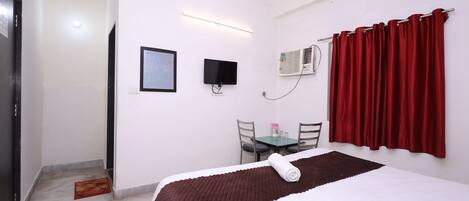 A hotel room with a bed, a desk, a chair, a TV, and an air conditioning unit.