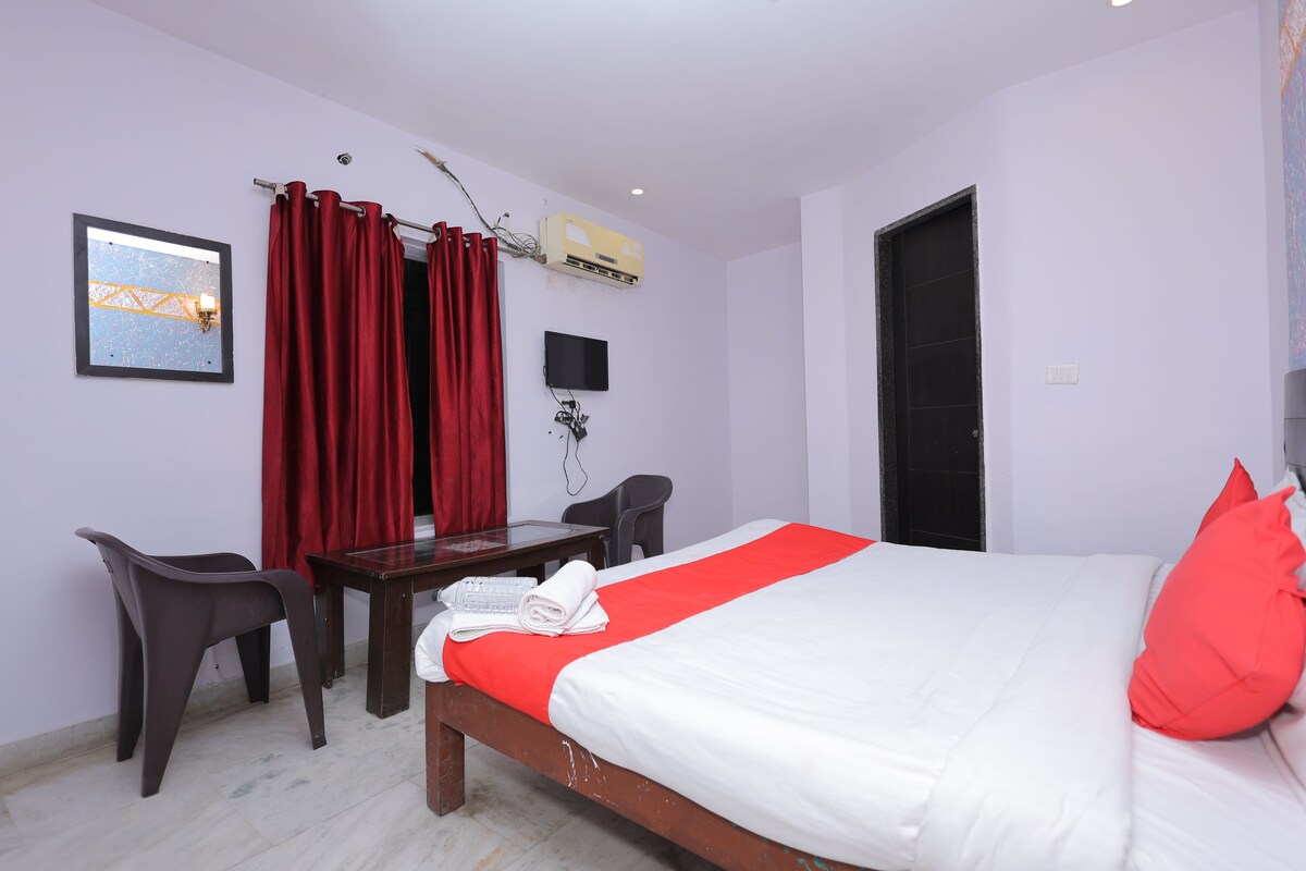 A hotel room with a bed, a desk, two chairs, a television, and an air conditioning unit.