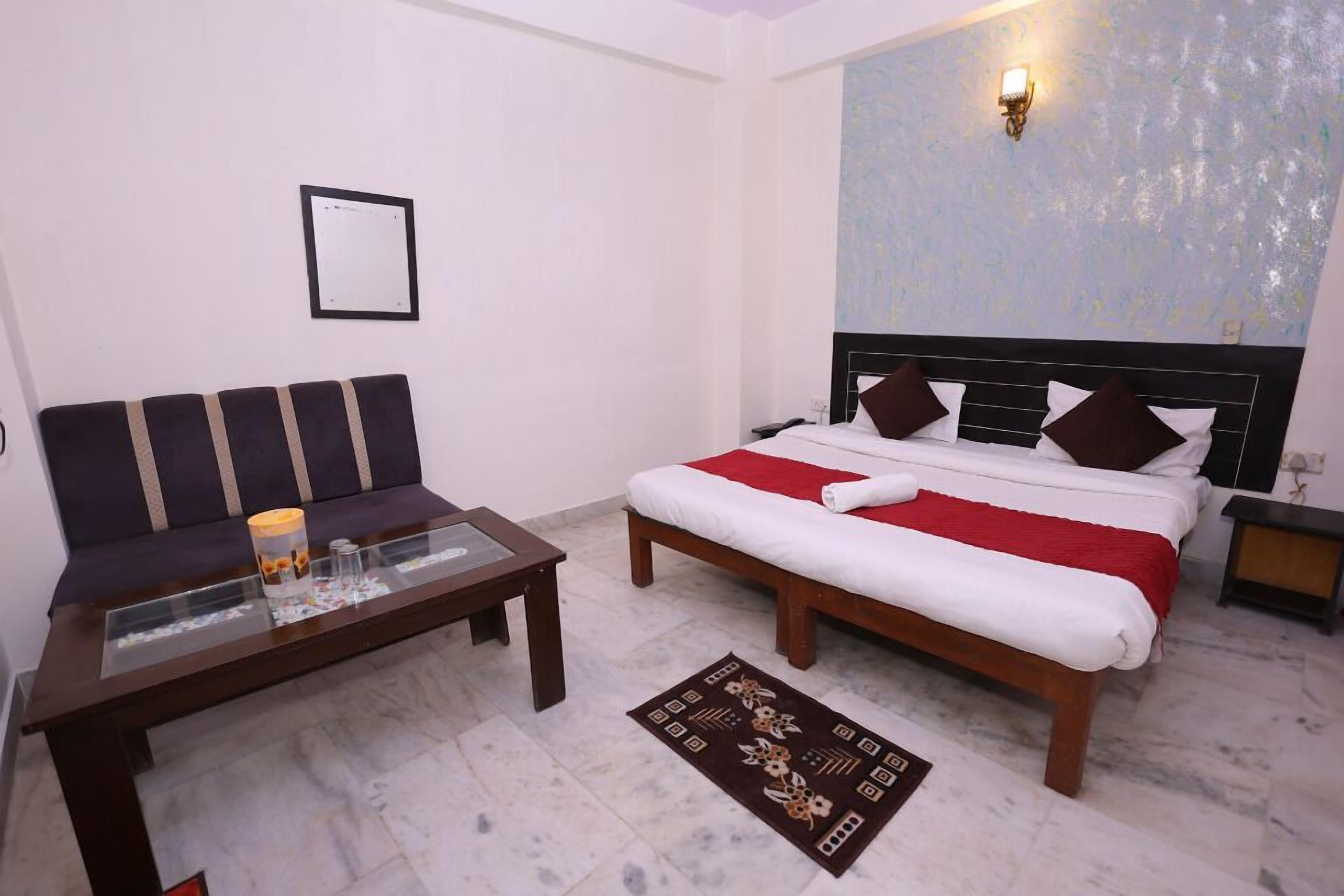 A hotel room with a bed, a sofa, a glass coffee table, a red and white bedspread, a patterned rug, a wall-mounted light, and a framed picture on the wall.