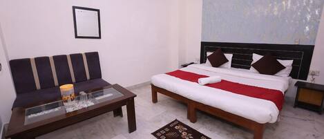 A hotel room with a bed, a sofa, a glass coffee table, a red and white bedspread, a patterned rug, a wall-mounted light, and a framed picture on the wall.