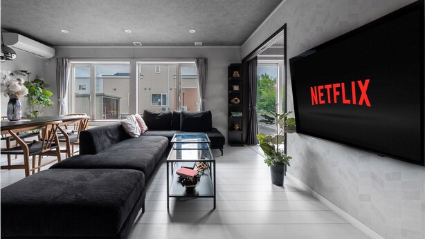 A modern living room with a large flat-screen TV displaying the Netflix logo, a black sofa, a glass coffee table, and a dining area with a table and chairs. - THE SHELBOURNE FURANO (Furano)
