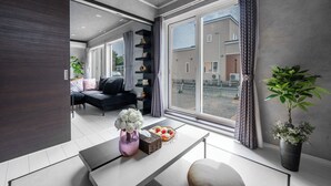 A modern living room with a sofa, a coffee table with flowers, and a large window with curtains. - THE SHELBOURNE FURANO (Furano)