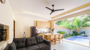 A modern living room with a sofa, a coffee table, and a dining area with a table and chairs. There is a kitchen with wooden cabinets and a refrigerator. A ceiling fan is visible, and a small pool is in the background. - Gold Fish House Ubud (Ubud)