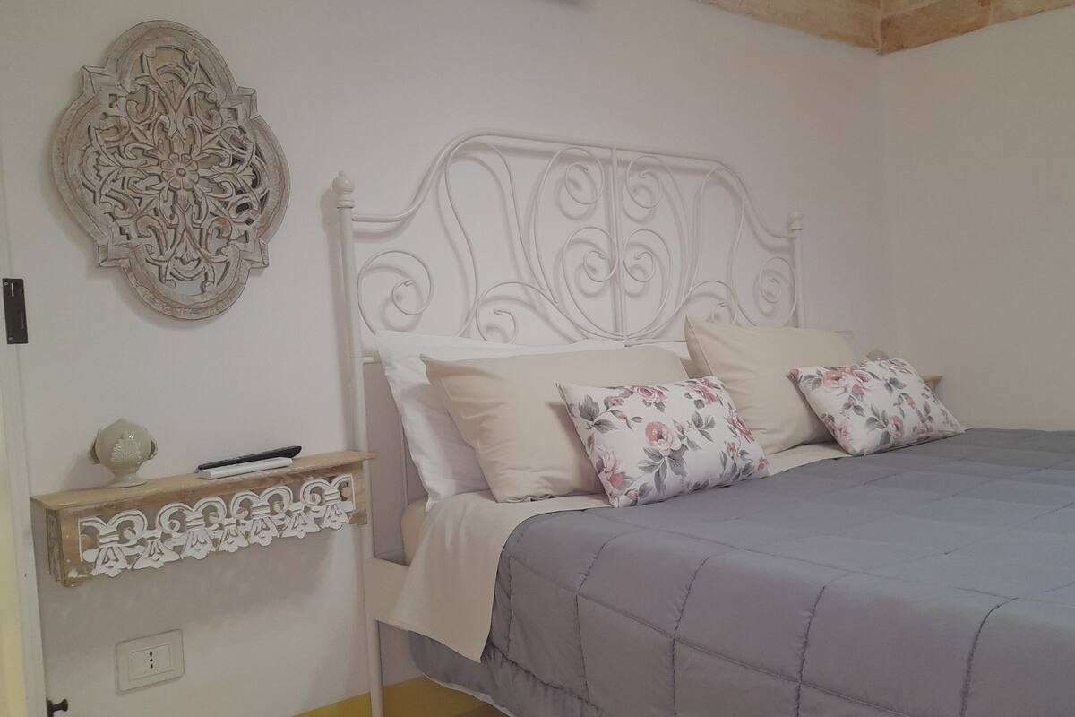 A bedroom with a white ornate headboard, a grey quilted bedspread, and floral-patterned pillows.