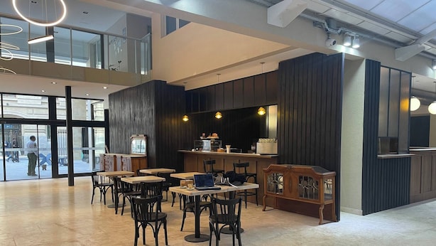 Un espace intérieur spacieux, doté de plusieurs tables et chaises, d’un comptoir avec un présentoir et d’une grande fenêtre donnant sur la rue.