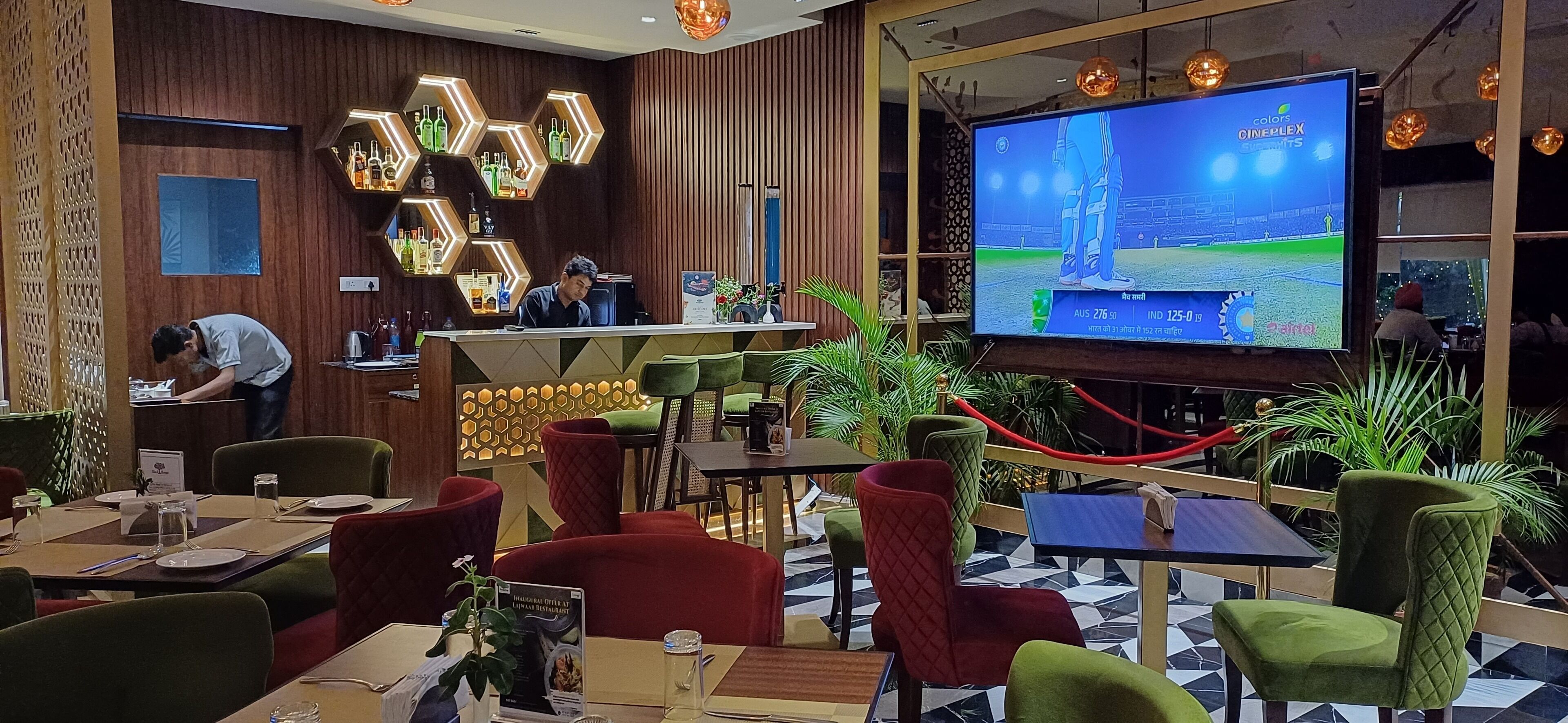 A restaurant with a bar area, a large screen displaying a cricket match, and various seating arrangements.