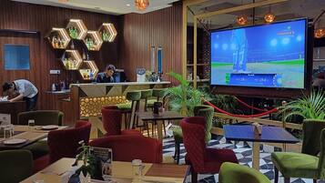 A restaurant with a bar area, a large screen displaying a cricket match, and various seating arrangements.