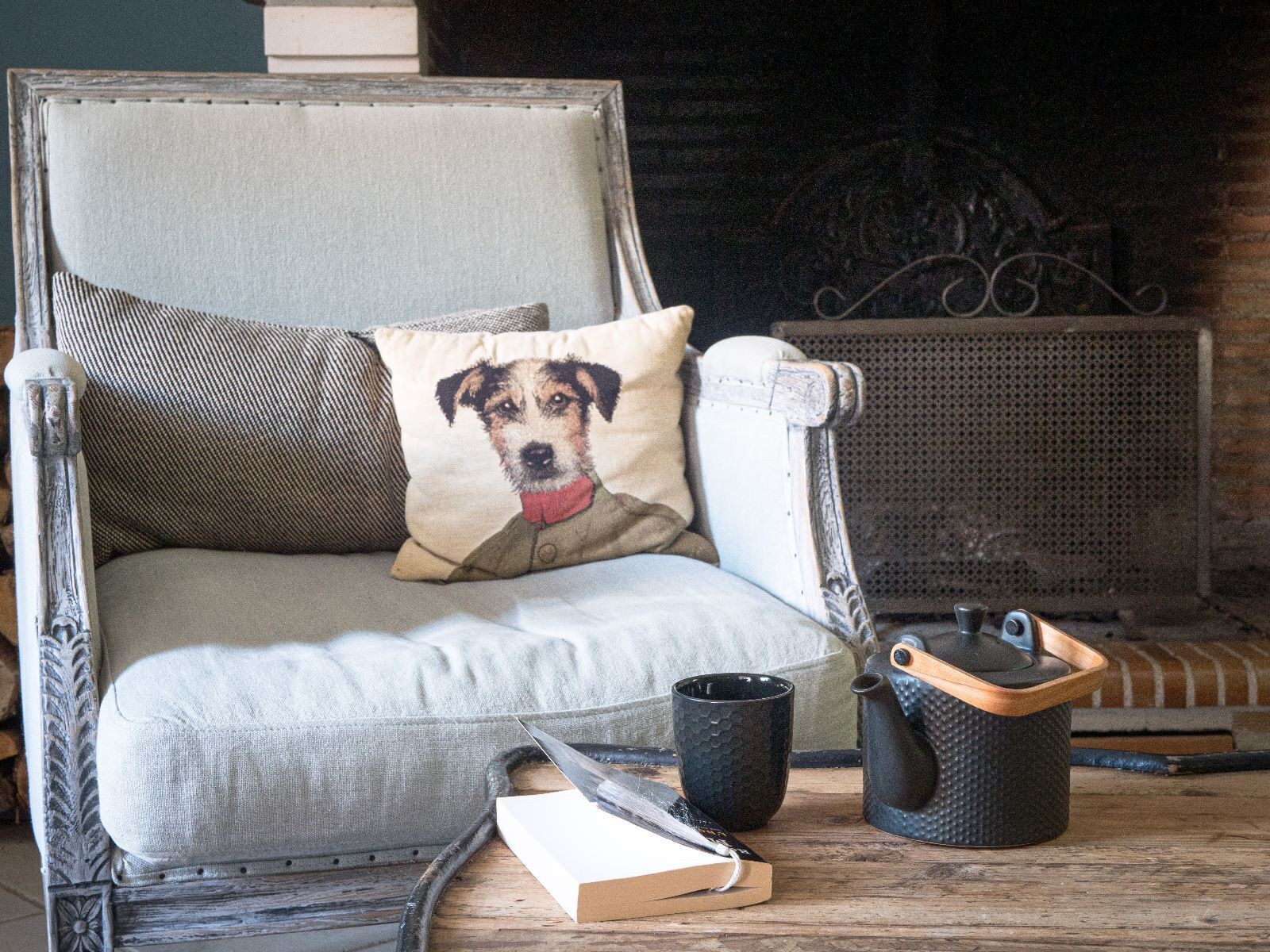 Una silla con un cojín que muestra un perro, un libro, una taza y un hervidor sobre una mesa de madera.