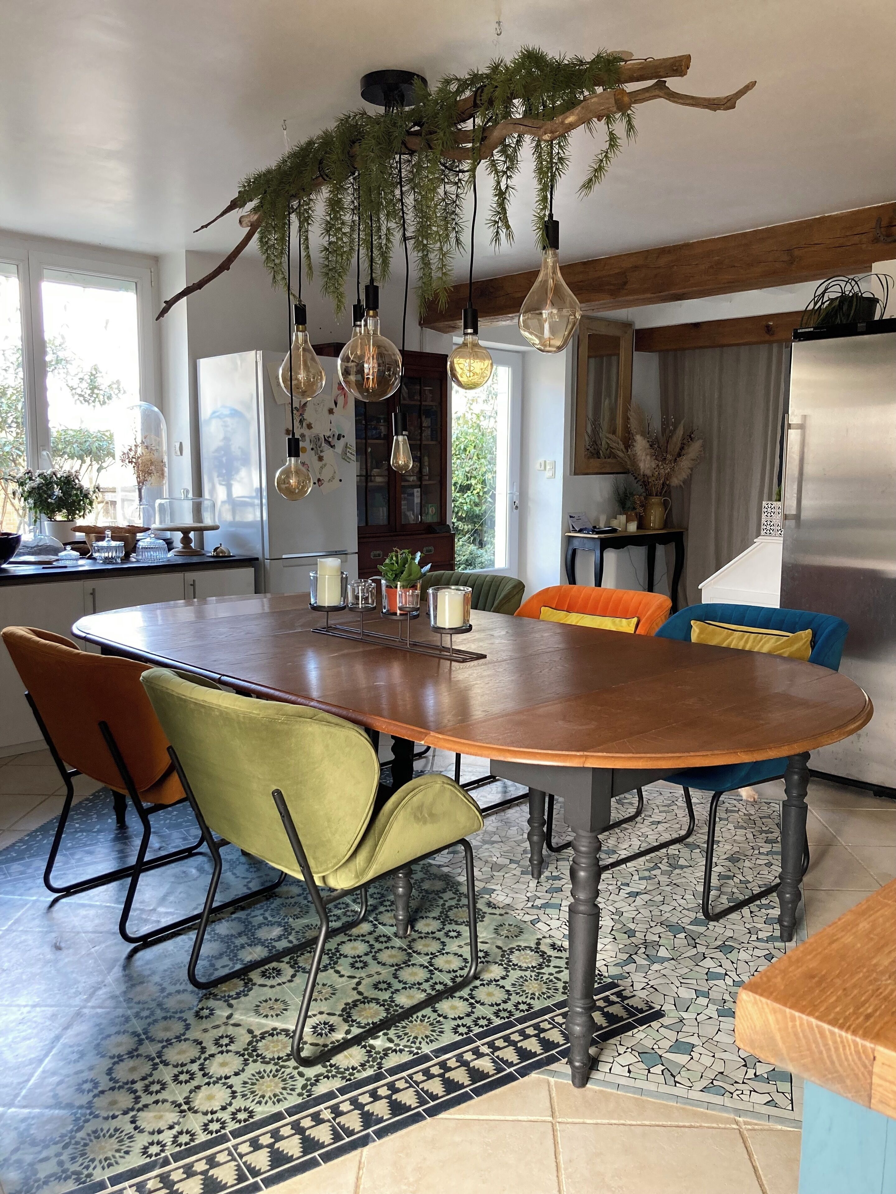 A modern kitchen with a large wooden table, colorful chairs, hanging light fixtures, and a decorative tree branch.