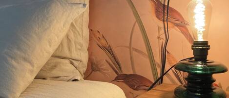 A bed with white bedding, a green lamp with a clear glass shade, and a wall with a floral mural.