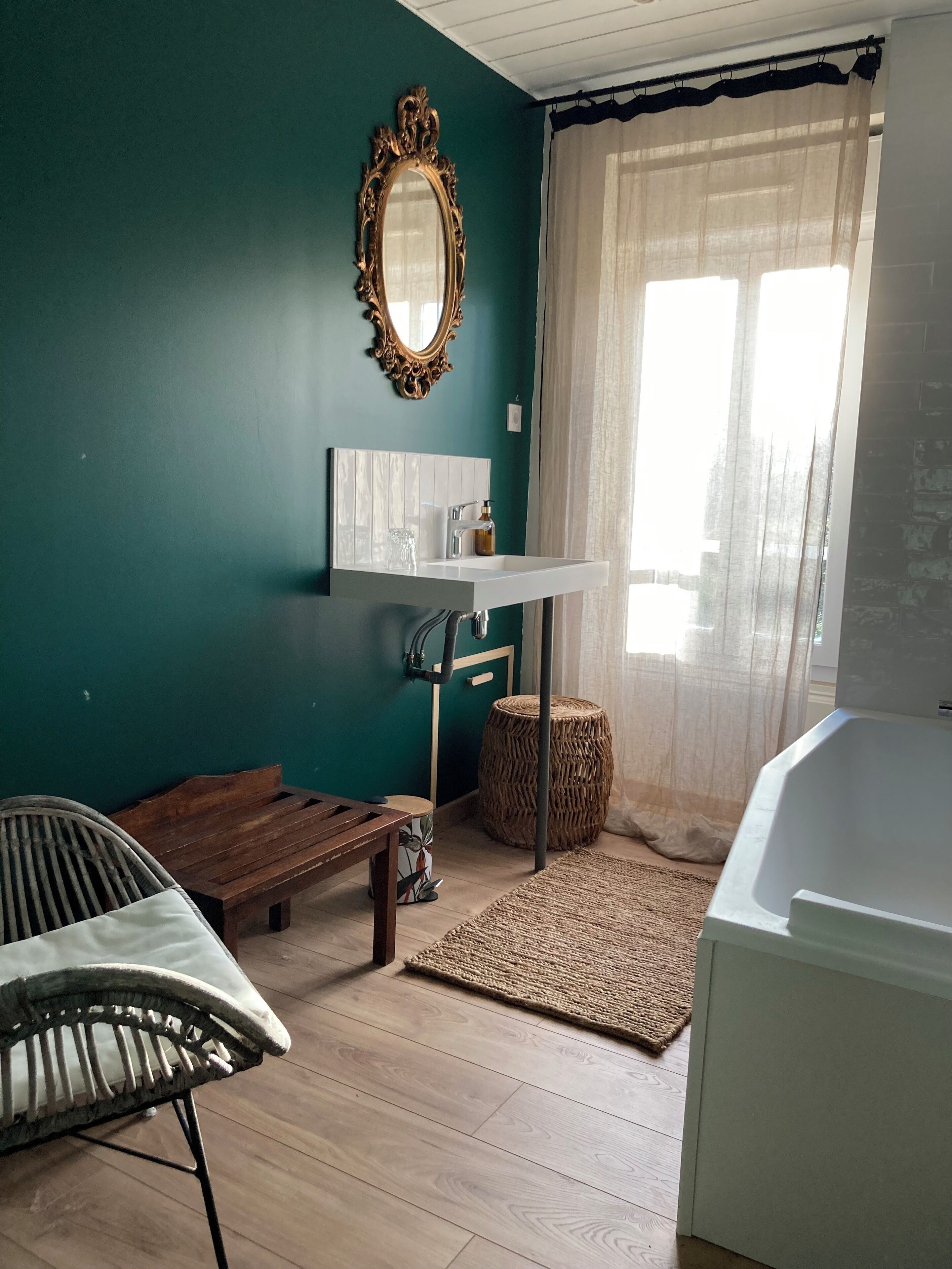 Een kamer met blauwgroene wanden, een houten bankje, een rieten kruk en een witte wastafel, met een groot raam met dunne gordijnen.