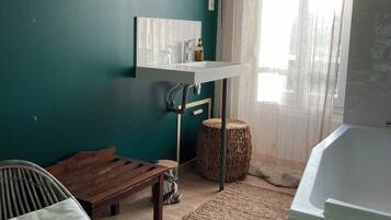 Een kamer met blauwgroene wanden, een houten bankje, een rieten kruk en een witte wastafel, met een groot raam met dunne gordijnen.