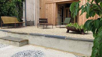 A wooden outdoor seating area with a bench and chairs, surrounded by gravel and plants.