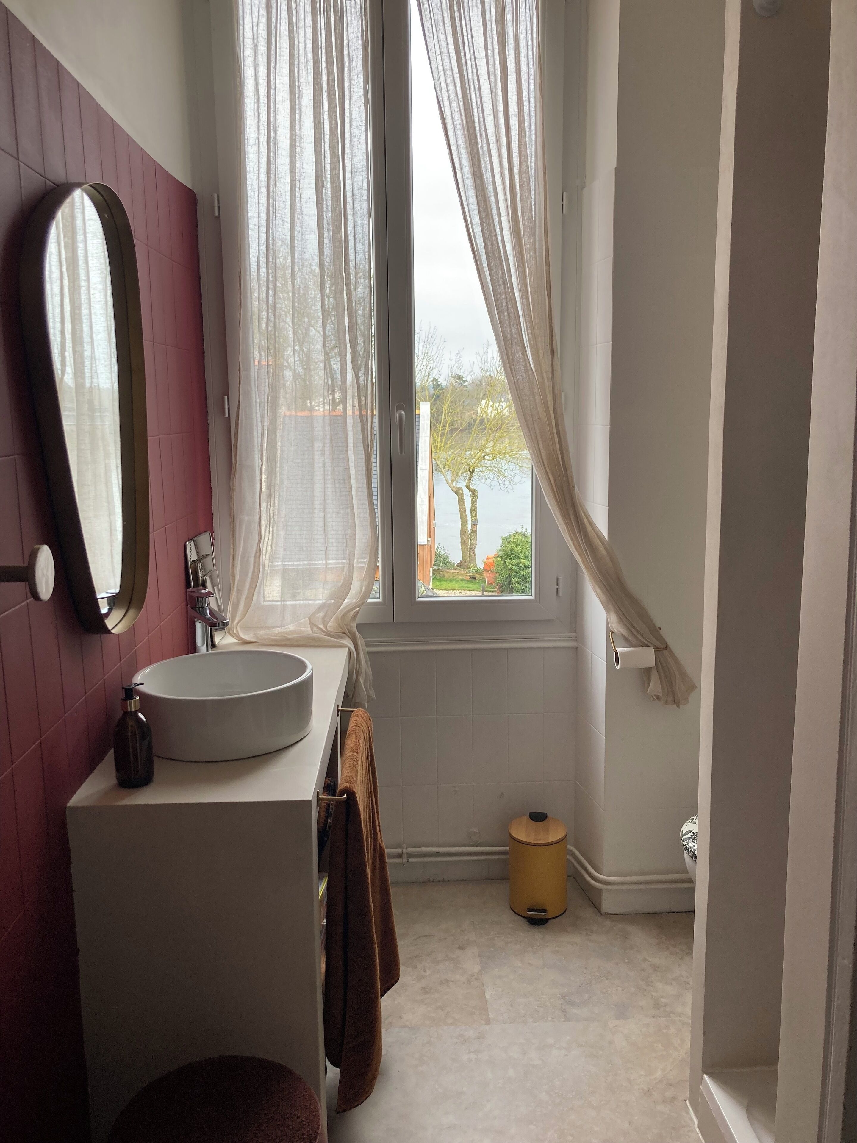 Ein Badezimmer mit einem weißen Waschbecken, einem Spiegel und einem Fenster mit transparenten Vorhängen.