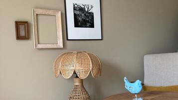 Una habitación con una mesa de madera, una lámpara de mimbre, una maleta y un adorno con forma de pájaro azul.
