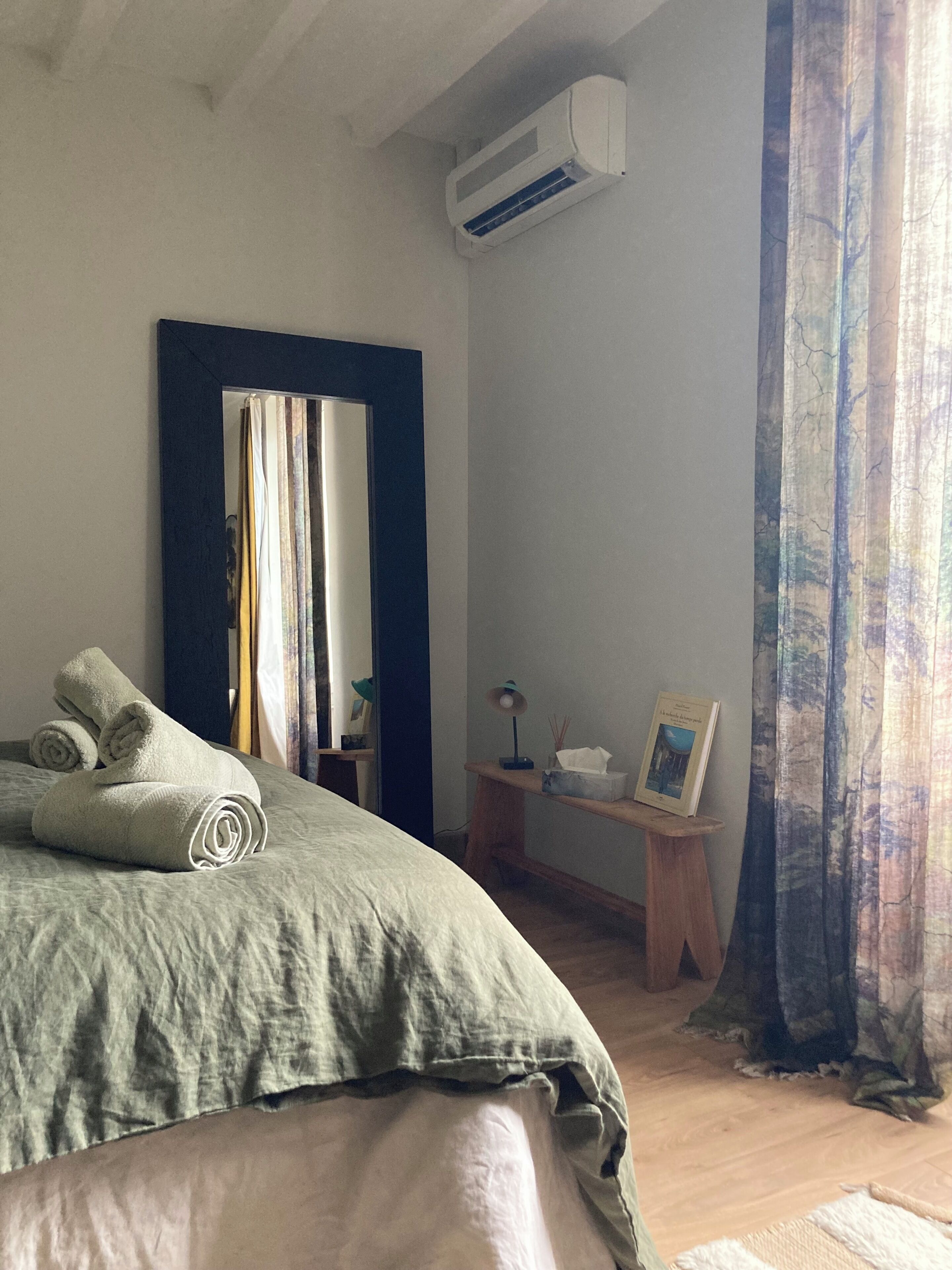 A bedroom with a bed, an air conditioner, a bedside table with a lamp, a picture frame, and a curtain.